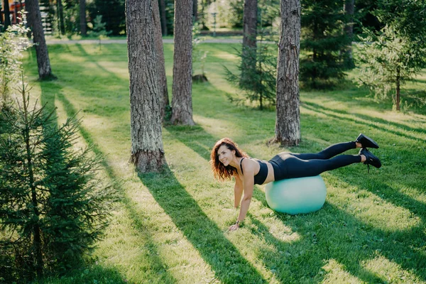 Determined young woman does Pilates in green park, balancing on fitness ball, wearing activewear, happily posing outside. Selective focus.