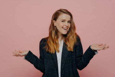 Orta boylu, kızıl saçlı, koyu renk ceketli ve beyaz bluzlu mutlu genç bir kadın. Pembe arka planda kapalı bir yerde el ele tutuşuyor. Vücut dili kavramı.