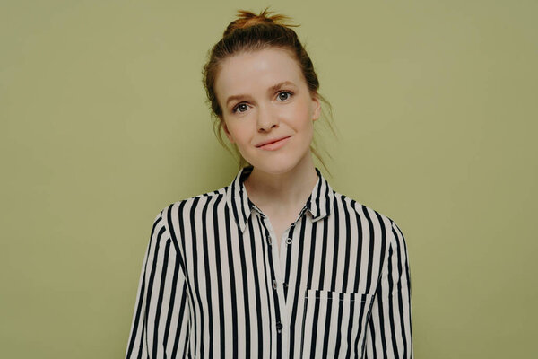 Calm young woman with bun, in black and white striped shirt, tilting head, slight smile, standing near green wall, looking at camera.