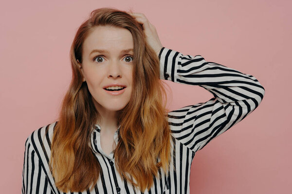 Attractive young woman in stripy black and white blouse, touching head in astonishment, expressing surprise against pink studio wall. Emotions concept.
