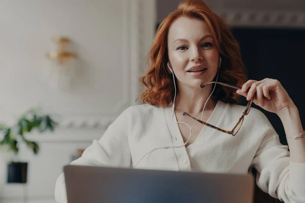準備された赤毛起業家 壮観とイヤホンを身に着けている 同僚とのオンライン交渉のための女性 不可欠な情報を検索するために彼女のラップトップを使用して ストック写真