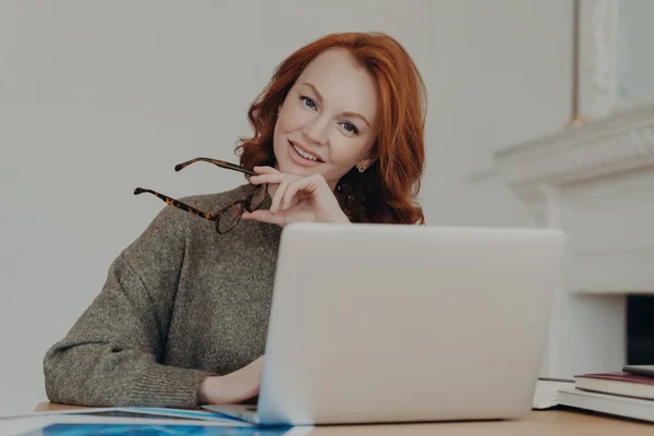 快乐的红头发学生利用她的笔记本电脑和互联网 在一个协作的空间里进行家庭作业研究 一个自由的网页设计师为客户设计 免版税图库图片