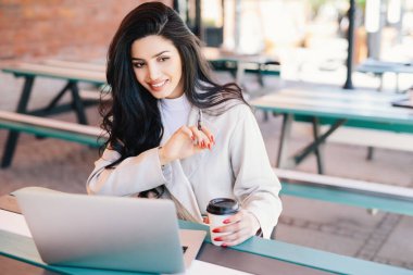 Açık kafede ücretsiz kablosuz internetin keyfini çıkaran, elinde kahve tutan ve genel bir dizüstü bilgisayar kullanan mutlu, serbest çalışan esmer kadın. Rahatlama, güzellik ve gençlik kavramlarını vurgulamak.