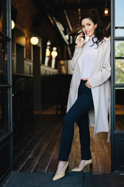 穿着深色头发 白色外套 黑色裤子和高跟鞋的时髦年轻女性 在咖啡店的背景下摆姿势 手挽手在手机前聊天 图库图片