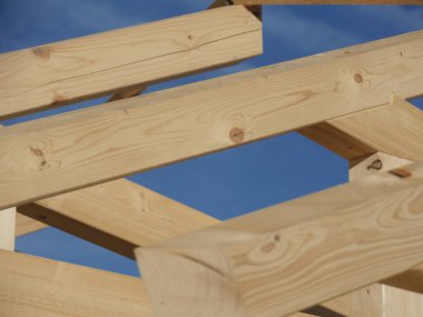 Tahta kirişler, yükseklik ve yapı hissi yaratacak şekilde dizilmiş. Arka plandaki mavi gökyüzü sahneye bir açıklık ve sakinlik hissi katar.