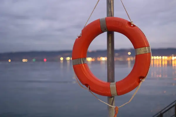 Kırmızı can yeleği metal bir direğe asılı. Su sakin ve gökyüzü bulutlu.