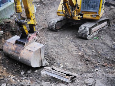 Sarı ve siyah bir inşaat makinesi çamura bir çukur kazıyor. Makine bir kepçedir ve hendek kazma aşamasındadır..