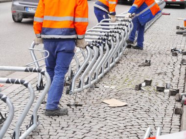 Turuncu ve mavi ceketli üç adam bir sıra metal askı üzerinde çalışıyor..