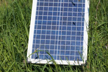 Bir güneş paneli çimlerin üzerinde oturuyor. Panel mavi ve beyaz. Çimler yeşil, gökyüzü mavi.