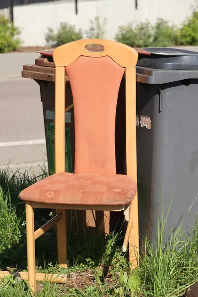 Kahverengi yastıklı ahşap bir sandalye bir çöp kutusunun yanındaki çimlerin üzerinde oturuyor. Sandalye eski ve yıpranmış görünüyor ve kullanılmıyor. Sahne terk edilme ya da ihmal edilme hissi veriyor.. .