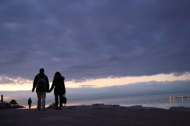 Gün batımında sahilde yürüyen bir çift. Gökyüzü bulutlu ve güneş batıyor. Çift el ele tutuşuyor ve suya doğru yürüyor.