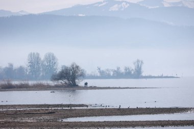 Bir çift deniz kenarında bir plajda yürüyorlar. Gökyüzü bulutlu ve su sakin.