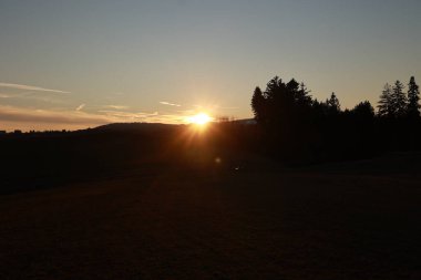 Arka planda ağaçlar olan bir tarlanın üzerinde güzel bir günbatımı. Gökyüzü turuncu ve pembe renklerin karışımıdır. Huzurlu ve huzurlu bir atmosfer yaratır.