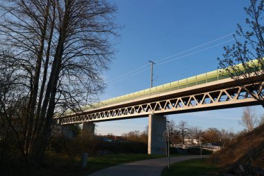 Altında tren akan bir nehir boyunca uzanan bir köprü. Köprü yeşil ve metal parmaklıkları var. Gökyüzü açık ve mavi, ve ağaçlar çıplak