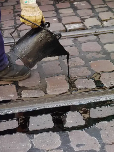 Adam bir kovaya siyah sıvı döküyor. Kova tuğla kaldırımda. Adam sarı eldiven giyiyor.