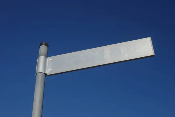 Mavi gökyüzünün önündeki bir direğin üzerinde sokak tabelası var. Tabela boş ve gümüş rengi var.