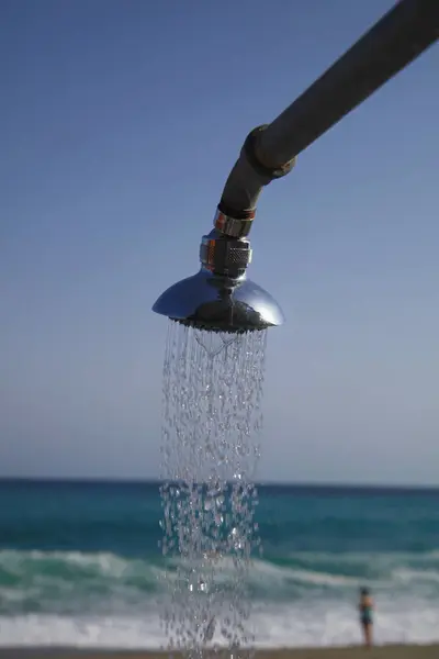 Duş başlığı havaya su püskürtüyor. Su sahile ve okyanusa dökülüyor. Sahne huzurlu ve rahatlatıcı.