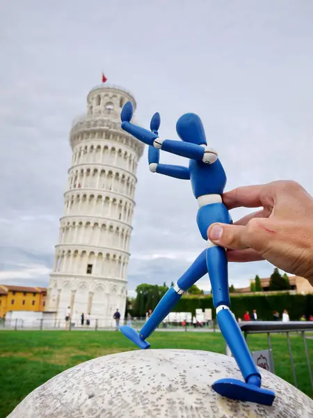 Mavi figür bir taşın üzerinde duruyor ve başka bir figürün elini tutuyor. Sahne Eğik Pisa Kulesi 'nin önünde geçiyor.
