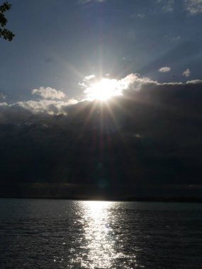 Güneş suyun üzerinde parlıyor, güzel bir yansıma yaratıyor. Gökyüzü bulutlu, ama güneş hala görünür, bir umut ve sıcaklık hissi yaratıyor.