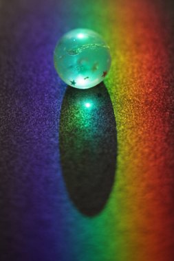 Küçük, parıldayan top karanlık bir yüzeyde oturur, gökkuşağı tarafından aydınlatılan bir gölge bırakır. Sahne huzurlu ve huzurlu, balo ve gölgesi huzur ve sükunet hissi yaratıyor.