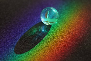 Gökkuşağı karanlık bir yüzeye yansıyor. Yüzeyde küçük, berrak, cam bir top duruyor.