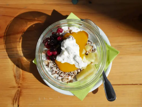 Masada bir kase meyve ve yoğurt. Kase muz, çilek ve yabanmersini de dahil olmak üzere çeşitli meyvelerle doludur. Yoğurdun üstünde tahıl gevreği ve bal var.