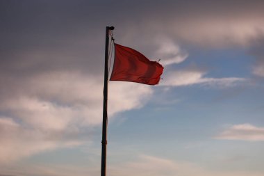 Gökyüzünde kırmızı bayrak dalgalanıyor. Gökyüzü bulutlu ve güneş batıyor. Bayrak biraz hasar görmüş, biraz da gözyaşı var.