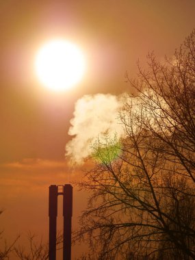 Gökyüzünde büyük, parlak bir güneş var ve ön planda büyük, siyah bir duman yığını var..