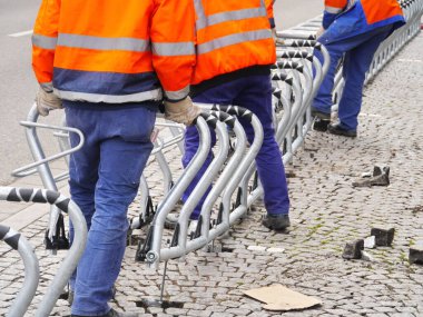Turuncu ve mavi güvenlik teçhizatlı üç adam metal bir çit üzerinde çalışıyor. Çit kırılmış ve tamir etmeye çalışıyorlar..