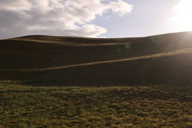 Bulutlu bir gökyüzü ve üzerinde güneş parlayan bir yamaç. Güneş çimlerin üzerine gölge düşürüyor