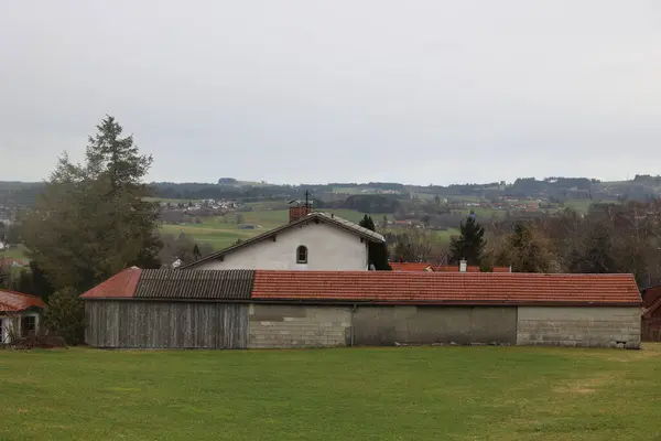 Kırmızı çatılı büyük bir ev çimenli bir tarlada oturur. Ev ağaçlarla çevrili ve bir bacası var. Gökyüzü bulutlu ve atmosfer huzurlu.