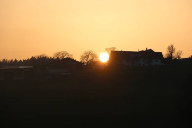 Bir evin penceresine gün batımı yansıtılır. Ev ağaçlarla çevrili ve gökyüzü turuncu ve pembe.