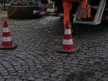Turuncu tulumlu bir adam iki kırmızı ve beyaz trafik konisinin önünde duruyor. Sahne bir şehirde geçiyor ve adam muhtemelen bir inşaat işçisi ya da trafik polisi.