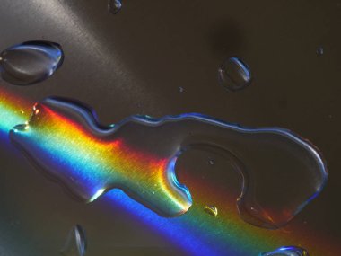 Gökkuşağı su birikintisine yansıyor. Su damlacıkları yüzeyin her tarafına dağılmış, parıldayan bir etki yaratıyor. Gökkuşağı parlak ve renkli görünüyor, neşe katıyor.