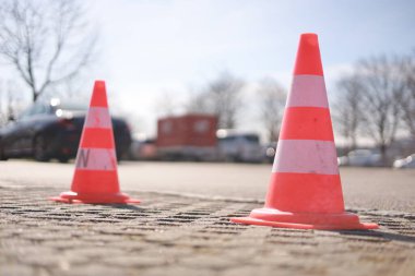 İki turuncu ve beyaz trafik konisi bir arabanın önünde yerde. Koniler bir otoparka yerleştiriliyor ve etraflarında park edilmiş birkaç araba var.