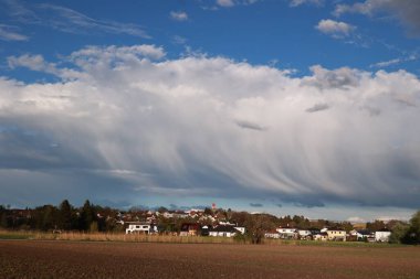 Gökyüzünde küçük bir kasabanın üzerinde büyük bir bulut var. Gökyüzü çoğunlukla mavi ve birkaç bulut etrafa saçılmış.
