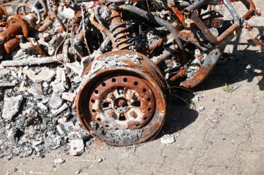 Bir araba, yanmış ve paslanmış bir tekerlekle bir hurdalıkta. Araba molozlarla çevrili ve arabanın tek görünen kısmı tekerlek. Sahne yıkım ve çürümenin bir parçasıdır.