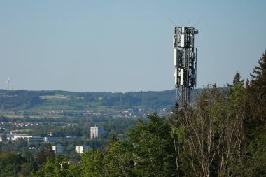 Üzerinde bir sürü anten olan yüksek bir kule. Kule bir ormanın ortasında ve arka planda bazı evler var. Gökyüzü açık ve güneş parlıyor.