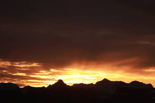 Güneş bir dağ sırasının arkasında batıyor, manzaranın üzerine sıcak bir parıltı saçıyor. Gökyüzü karanlık ve bulutlu, arkaplanda dağlar belirdi. Sahne huzurlu ve huzurlu.