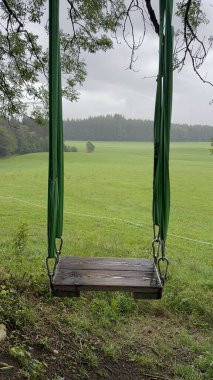Yumuşak bulutlu bir gökyüzünün altında yemyeşil bir manzarayla çevrili, ağaçlardan salınan sakin bir salıncak.
