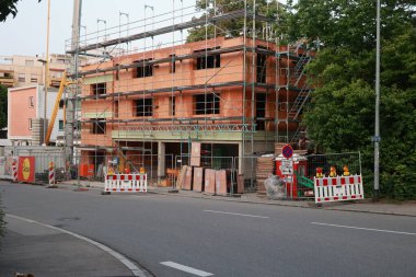 İnşaat alanı bir caddenin kenarında görünür. Bina inşa ediliyor ve şantiyede çok sayıda işçi var. İşçiler turuncu yelek giyiyor ve bir sürü turuncu koni var.