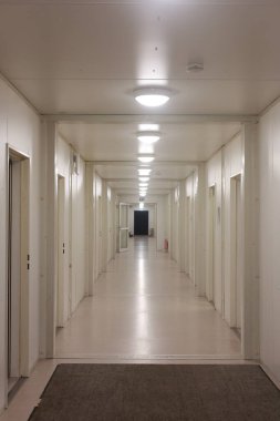 Beyaz duvarları ve kapıları olan uzun bir koridor. Koridor boş ve hiç insan yok. Işıklar açık ve koridor aydınlık.