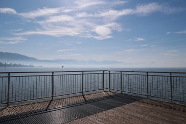 Büyük bir su kütlesine bakan korkuluklu ahşap bir güverte. Gökyüzü bulutlu, sakin bir atmosfer yaratıyor. Suyun manzarası nefes kesici ve güverte rahatlamak için mükemmel bir yer.