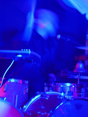 Adam bir grupta davul çalıyor. Davullar mavi ve adam siyah bir ceket giyiyor.