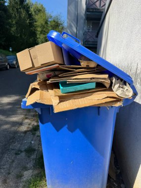 Bir yerleşim bölgesinde bulunan mavi bir geri dönüşüm kutusu karton ve kağıt atığı bolluğu ile doludur.