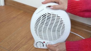 Kadın elinde kırmızı bir kazakla odada elektrikli vantilatör ısıtıcısı ayarlıyor ve sıcak olup olmadığını eliyle kontrol ediyor. Kız odayı ısıtmak için taşınabilir pervane ısıtıcısını açıyor. Özerk elektrik alan ısıtması