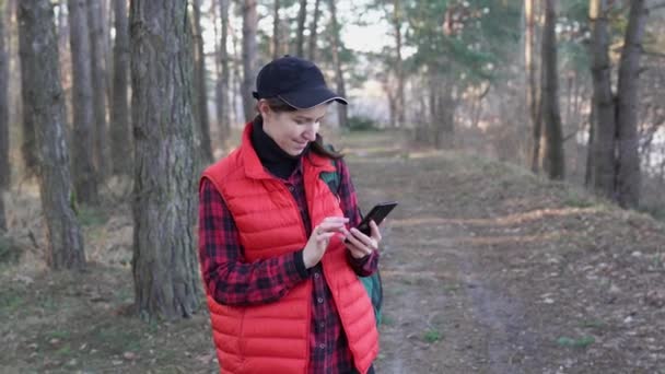 森の中のバックパック付きの赤いベストで幸せなハイキングの女の子は 電話でメッセージを入力しています ソロはスマートフォンを手に若い女性を旅する 松の木の間の女性ハイカーはガジェットを使用します — ストック動画