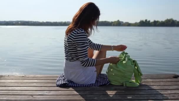 一头长发的女孩背靠着背坐在靠近水的木制码头上 在他的背包里寻找面具 年轻的黑发女子通过肩袋在河里或湖中的泛泛而谈 — 图库视频影像