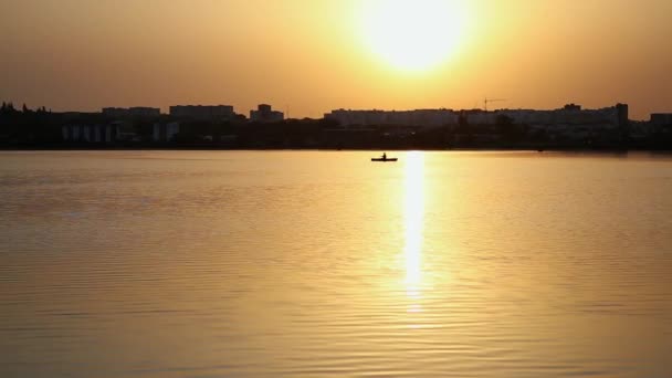 天気が静かで 穏やかな湖の漕ぎ耳の上に小さなボートセーリングボートで1人の男 都市の背景に風のない日の上昇の光線で川のボートで漁師の帆のシルエット — ストック動画