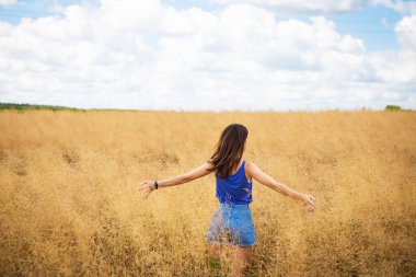 Mavi tişörtlü ve kot şortlu güzel esmer bir kadının portresi tarlanın ortasında duruyor, açık havada bir yaz gününün arka planında yüzünü güneşe gösteriyor.
